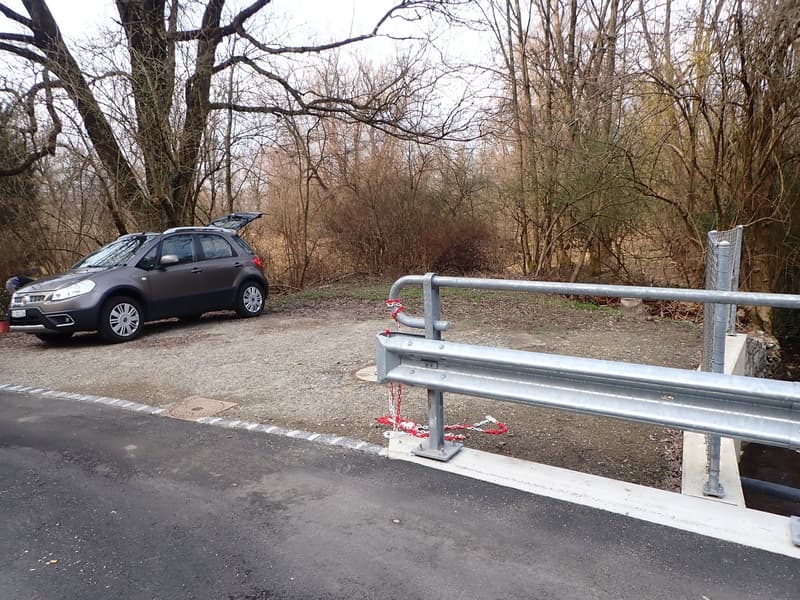 Outdoor parking area with a small car parked on gravel, surrounded by bare trees and a metal guardrail with chains.
