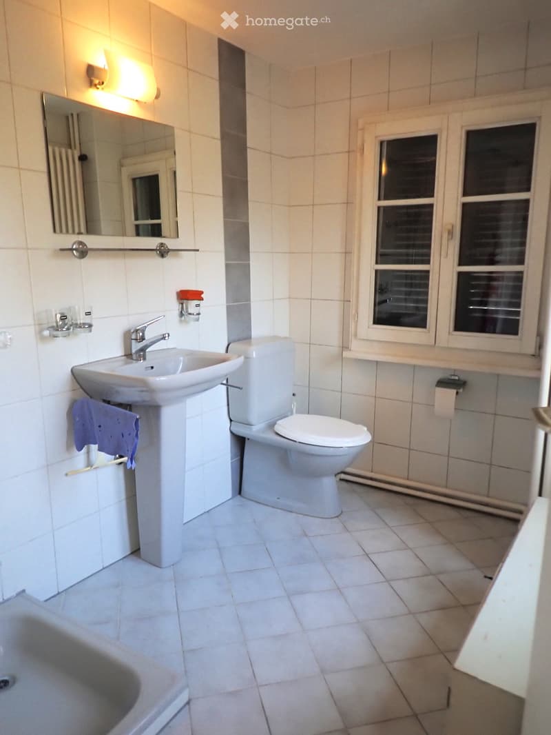 Clean, modern bathroom with white tiles, a sink, toilet, mirror, and a towel hanging on the wall.