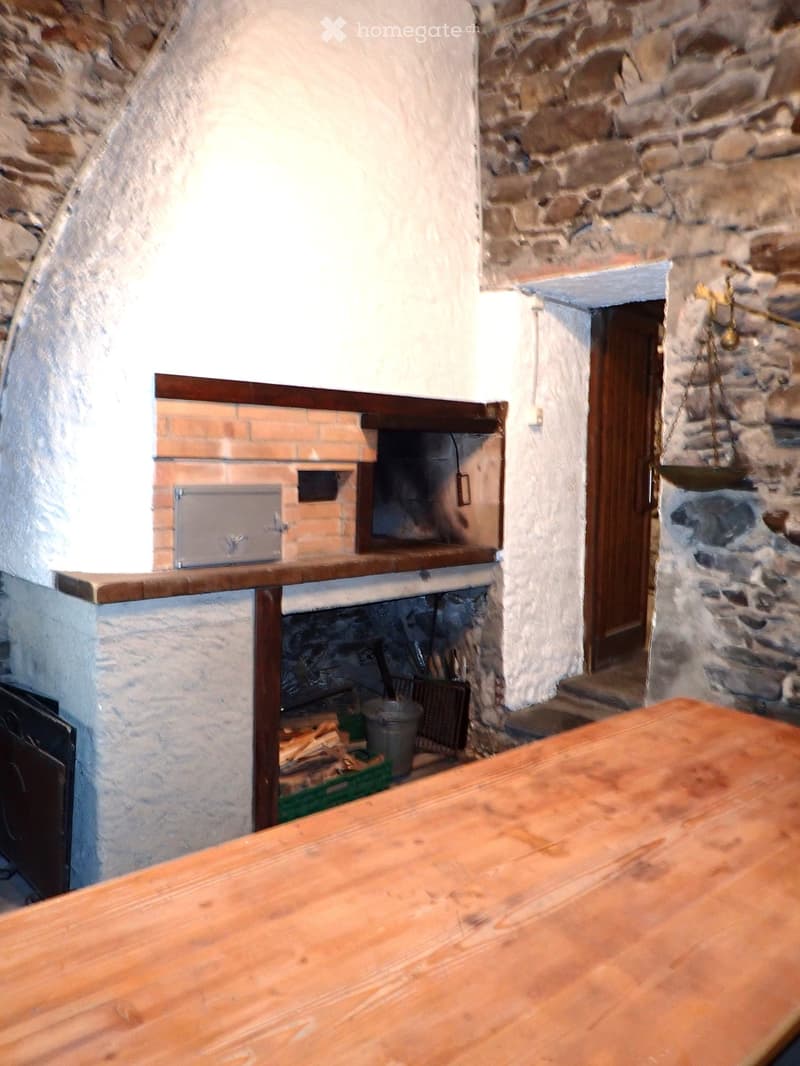 A rustic room with a large wooden table in the foreground, a stone fireplace with a wooden shelf above it, and a wooden scale hanging on the wall. The room features exposed stone walls and a wooden door leading to another area.