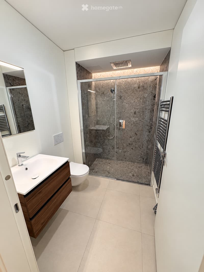 Modern bathroom with a white sink, toilet, glass-enclosed shower, and a large mirror. The floor is tiled in light beige, and the walls are white with a dark, textured shower area.