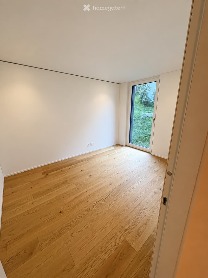 Empty room with light wooden flooring, white walls, and a large sliding glass door leading to a green outdoor area.
