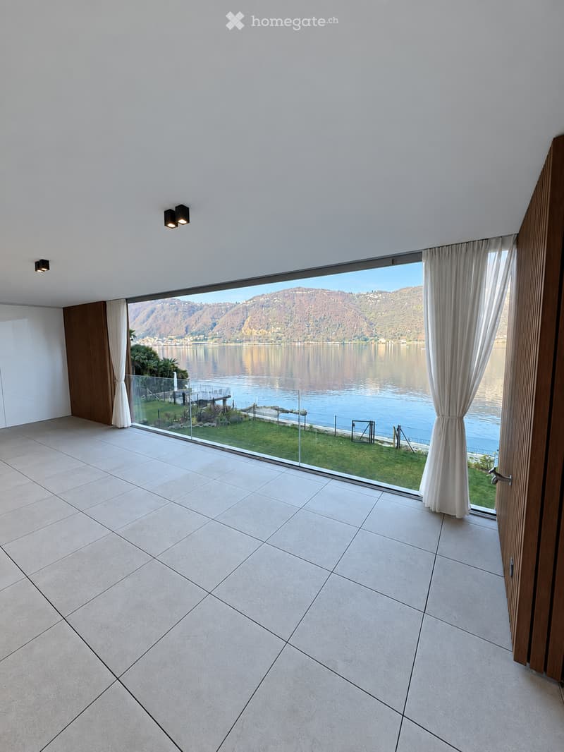 Empty room with large glass windows offering a scenic lake and mountain view, tiled floor, and white walls.
