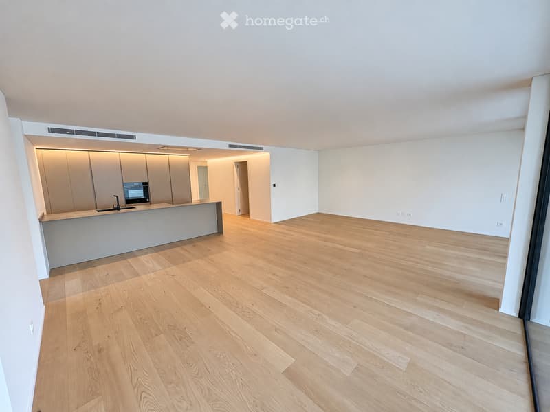 Empty room with light wooden flooring and white walls, featuring a modern kitchen area with cabinets and a countertop in the background.