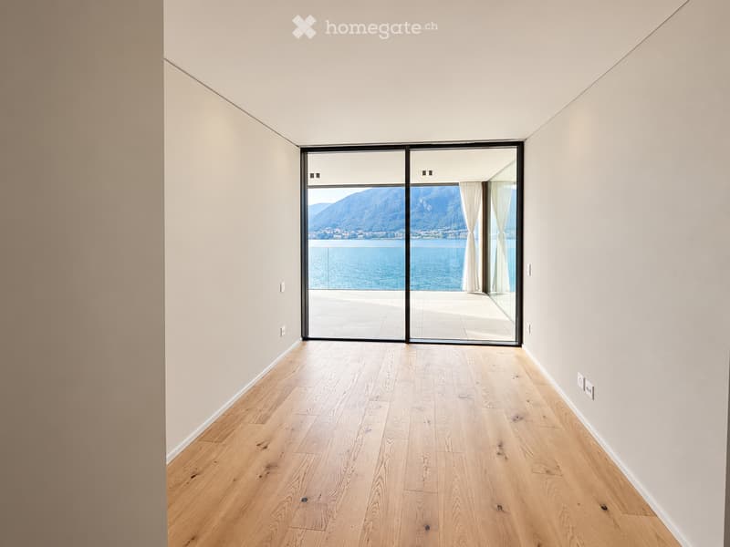 Empty room with light wooden flooring and white walls, featuring large sliding glass doors that open to a balcony overlooking a scenic view of mountains and water.