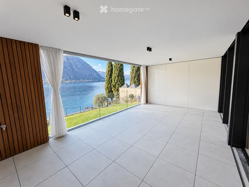 Empty room with large windows offering a scenic view of a lake and mountains, white tiled floor, and modern lighting fixtures.