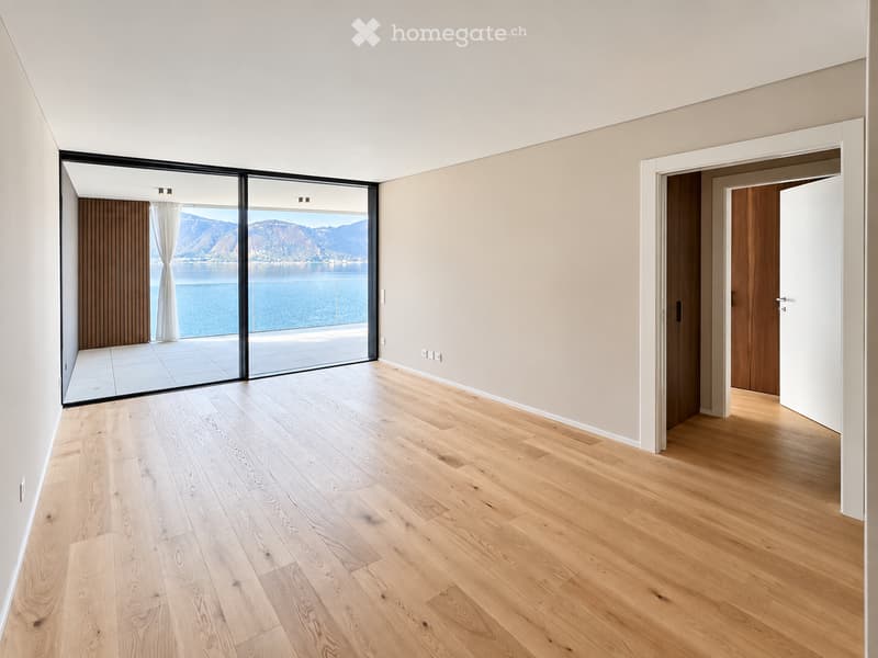 Empty room with light wooden flooring, white walls, large glass sliding doors leading to a balcony with a scenic lake and mountain view.