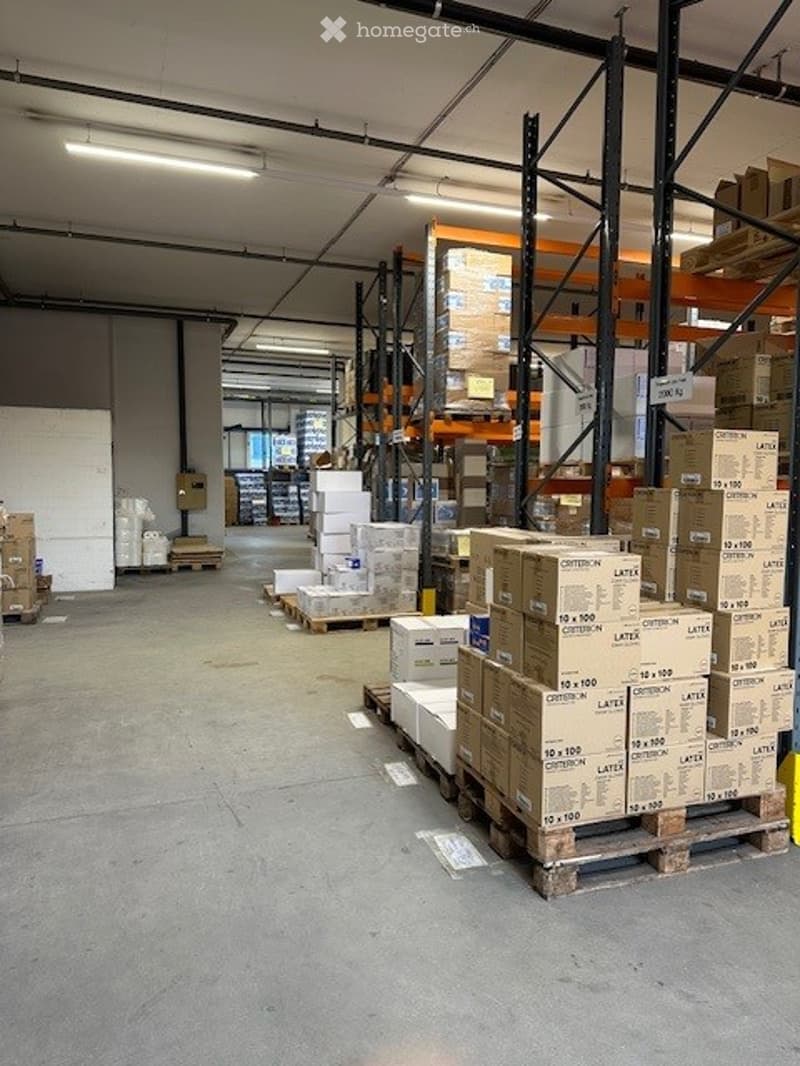 A large warehouse with a high ceiling, fluorescent lights, pallets stacked with various boxes, white walls, and an aisle with forklifts