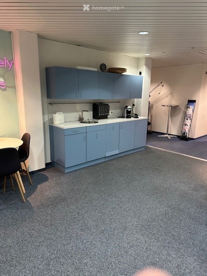 A room with a kitchen setup that includes a sink, cabinet, and coffee maker. The walls are painted white, and there is a blue carpet. A desk and chairs are present in the room, along with a microwave.