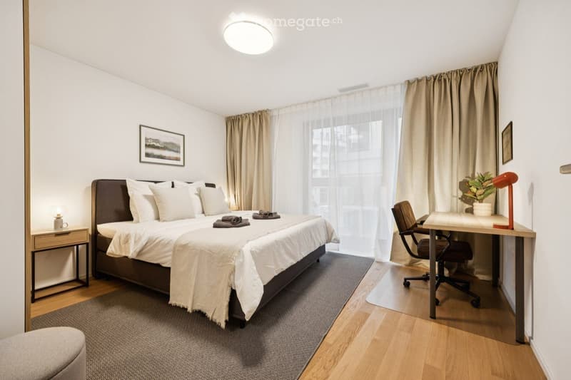 Modern bedroom with a large bed, nightstand, desk, chair, and decorative elements including a plant and artwork.