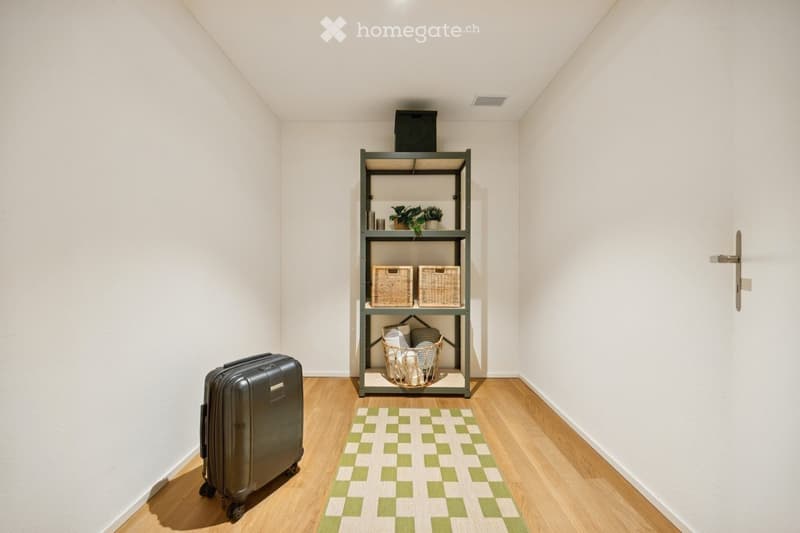 A small, neatly arranged room with light-colored walls and wooden flooring. A metal shelf holds baskets, plants, and other small items. A patterned rug lies on the floor, and a large suitcase is positioned near the wall.