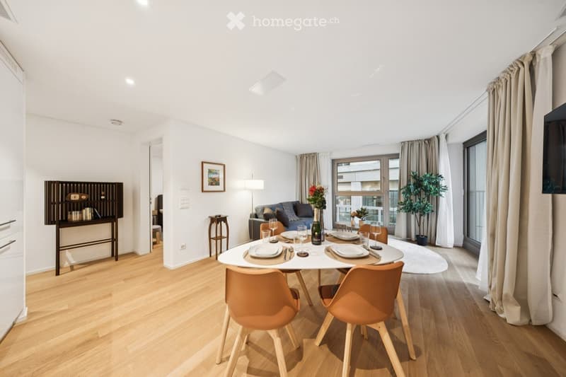 Modern, well-lit living and dining area with wooden flooring, white walls, and large windows. Features a sofa, dining table with chairs, a small cabinet, a table lamp, and decorative plants.