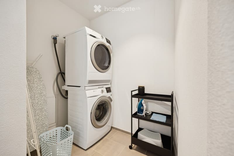 A small, well-organized laundry room with a stacked washer and dryer, an ironing board, and a shelf holding various laundry items.