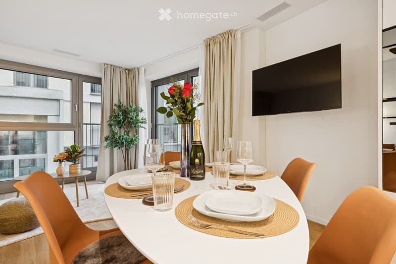 Dining area with a white table set for six, surrounded by orange chairs, a bottle of champagne, and decorative vases with flowers. Large windows offer a view of the city.