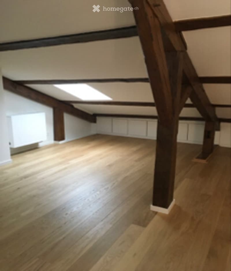 An empty attic space with exposed wooden beams and a skylight, featuring a polished wooden floor and white walls.