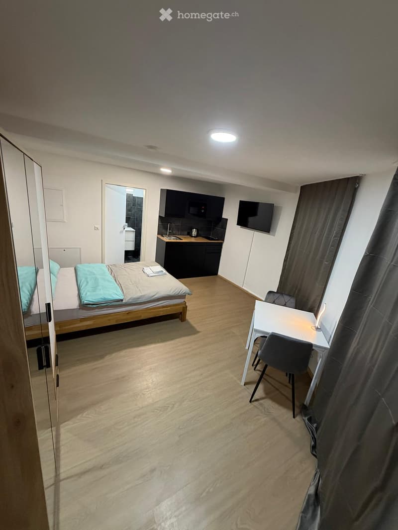 A compact, furnished room with a bed, a small kitchen area, a desk with a chair, and a television mounted on the wall.