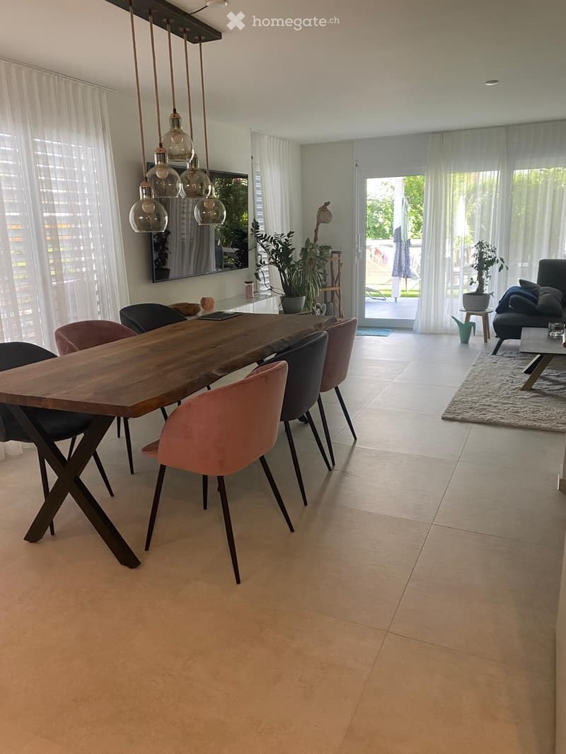 Modern open-plan living and dining area with a large wooden dining table, colorful chairs, a sofa, coffee table, and large windows with sheer curtains.