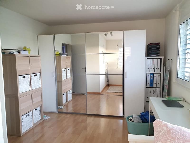 A well-lit room with wooden flooring, a large mirrored wardrobe, a storage unit with baskets, a desk with a laptop, and a small chair.