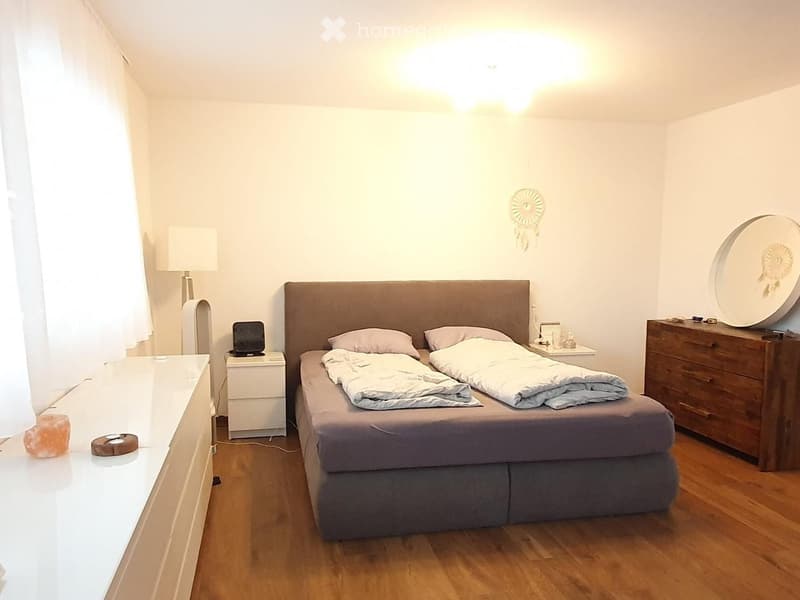 A cozy bedroom with a large bed, nightstand, lamp, and a wooden dresser. The room features wooden flooring and white walls.
