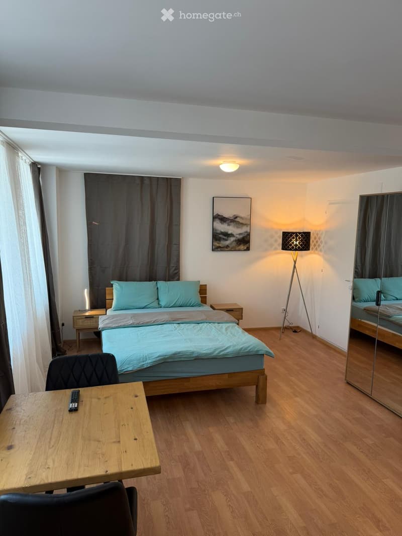 Cozy bedroom with a wooden bed, nightstands, a wooden table with a chair, a floor lamp, and a mirror. The room features light blue bedding and a scenic picture on the wall.