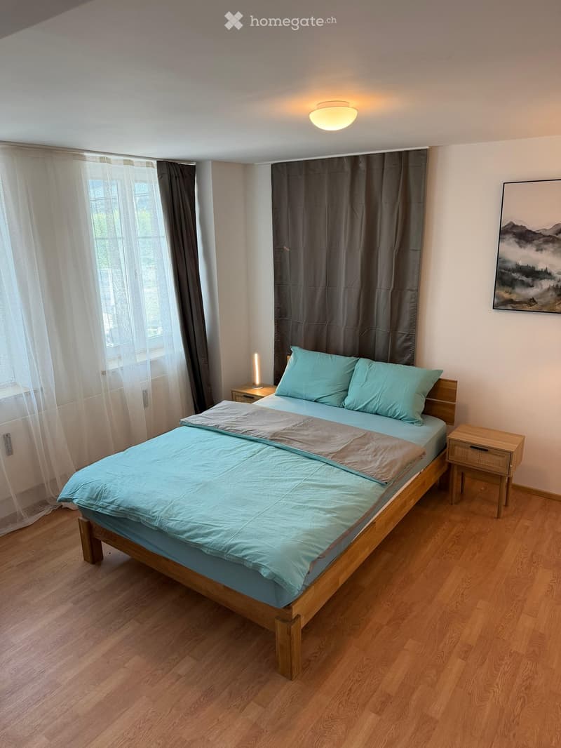 Cozy bedroom with a wooden bed, light blue bedding, nightstands, and a large window with sheer curtains.
