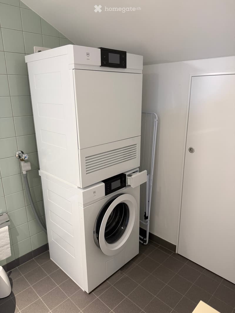 Modern bathroom with a stacked white washing machine and dryer unit, tiled walls, and a door.