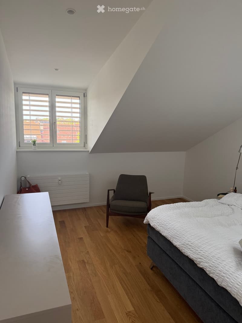 Cozy attic bedroom with wooden flooring, a large window with blinds, a bed with white bedding, a grey armchair, a radiator, and a small table with a handbag.