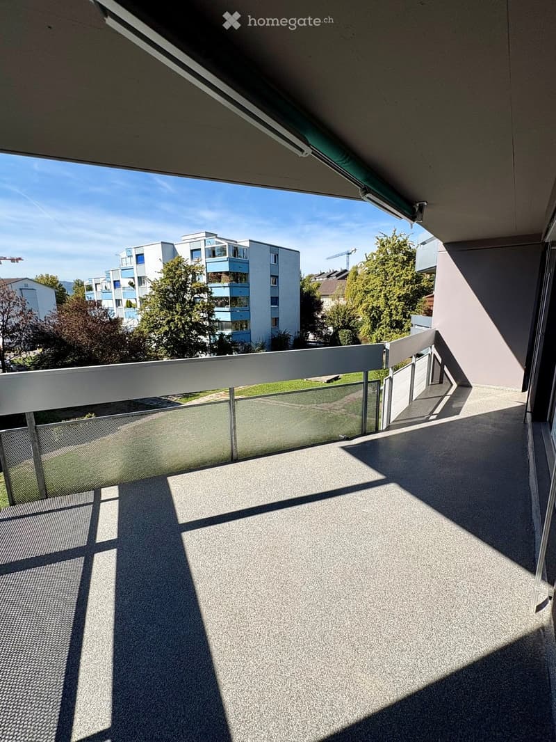 Balcony with modern glass railing, concrete floor, and view of surrounding buildings and greenery.