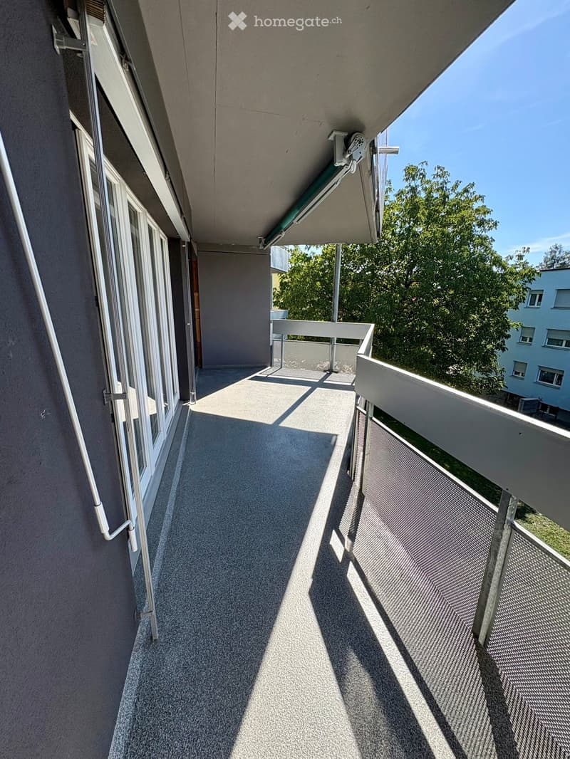 Balcony with grey carpeted floor, white metal railing, and large windows, overlooking a green area with trees and neighboring buildings.