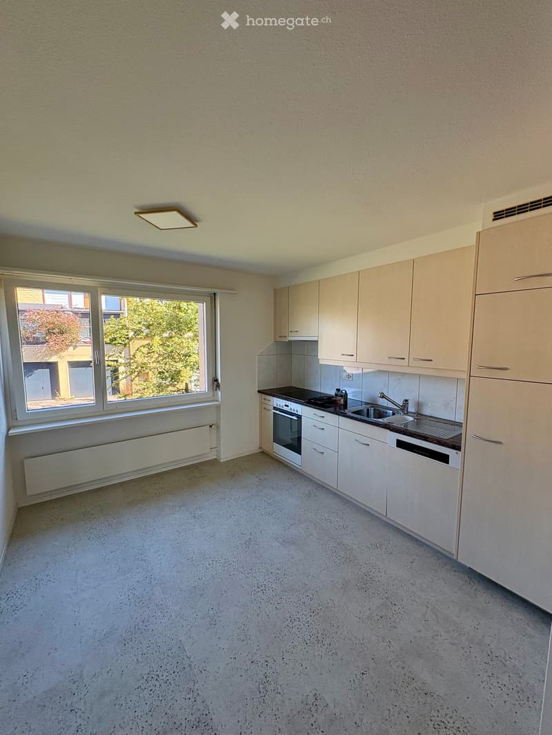 Empty kitchen with white cabinets, a sink, and an oven, featuring a large window with a view of greenery outside.
