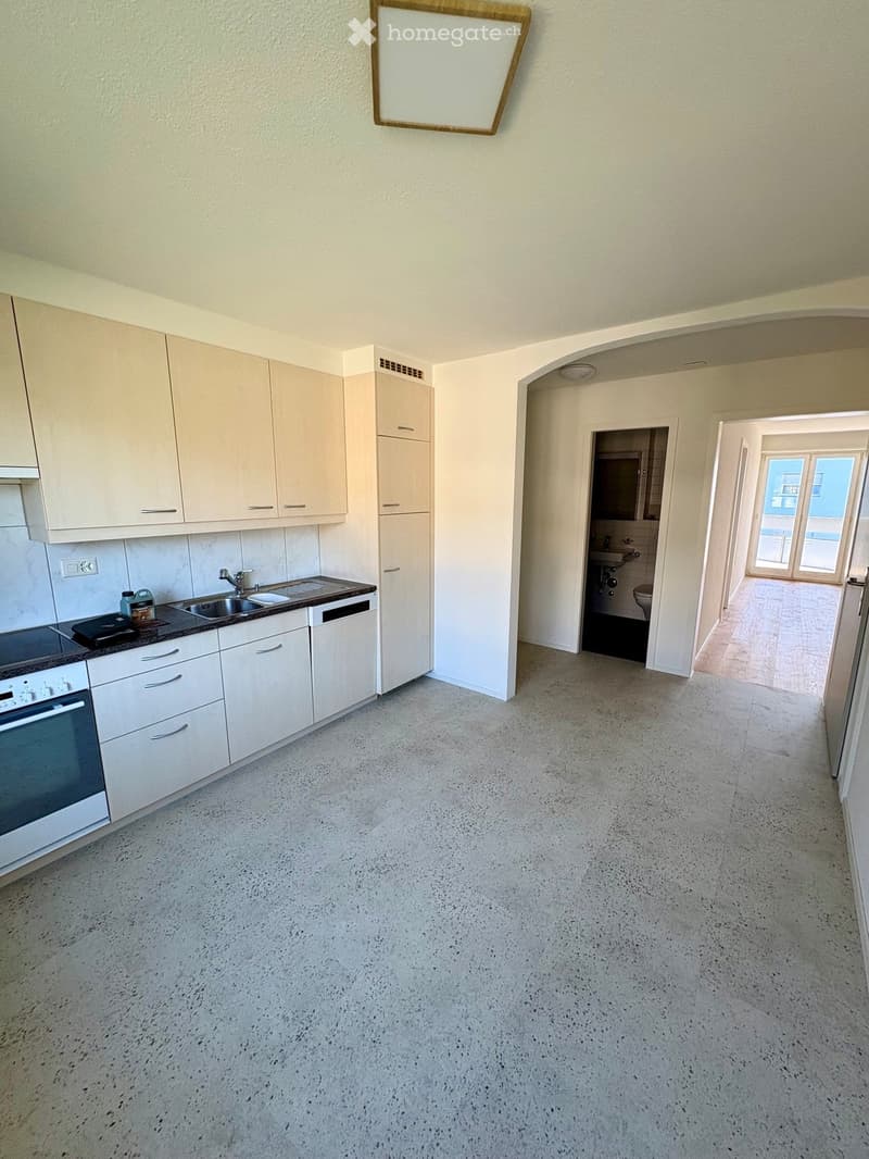 Empty kitchen and hallway area with white cabinets, sink, and a view into a small bathroom. The floor is concrete, and the walls are light-colored.
