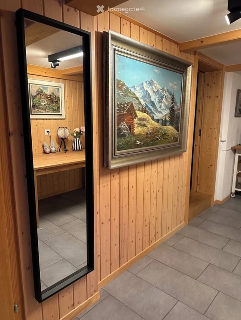 A small, well-lit room with wooden panel walls and a large mirror reflecting a painting of a mountain landscape. A wooden table holds a vase with flowers and a lamp, with additional artwork visible in the background.