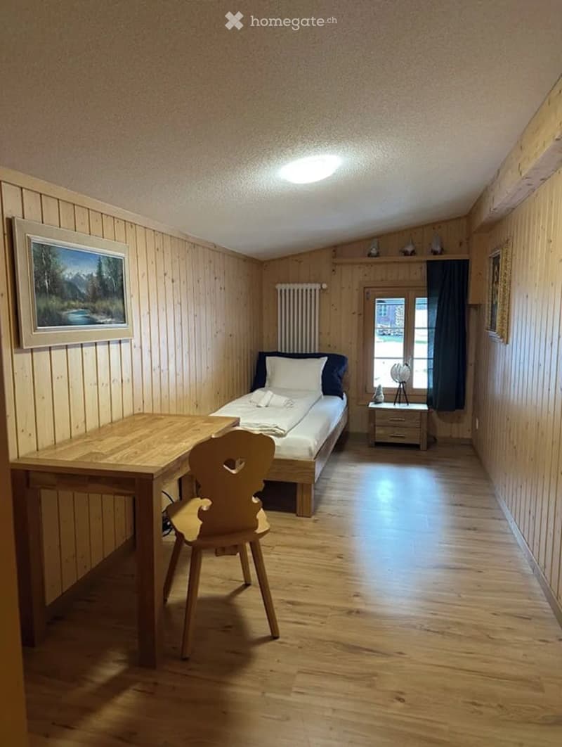 Cozy wooden-paneled bedroom with a single bed, desk, chair, nightstand, and a window with dark curtains.