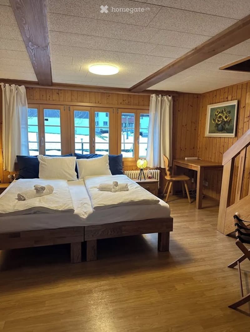 Cozy wooden bedroom with two single beds, nightstands, a small desk, and a chair. Large windows with curtains let in natural light.