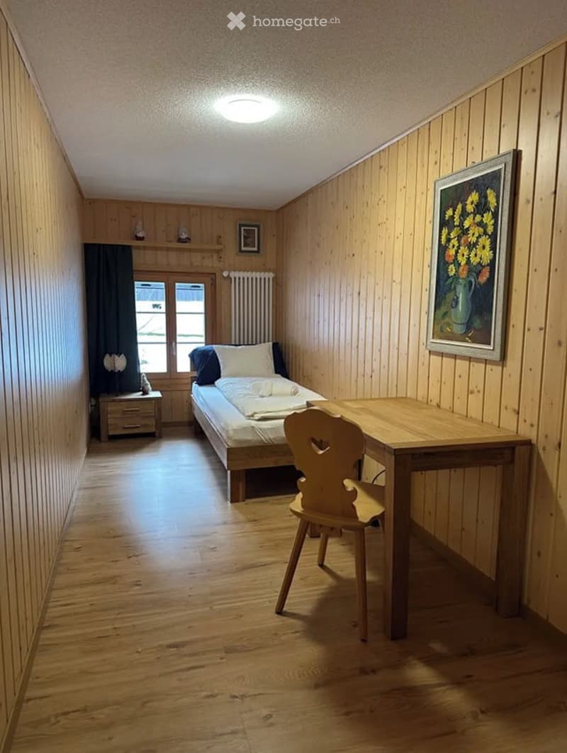 Cozy bedroom with wooden walls and floor, single bed with white and blue bedding, nightstand, wooden chair, small table, and framed artwork on the wall.