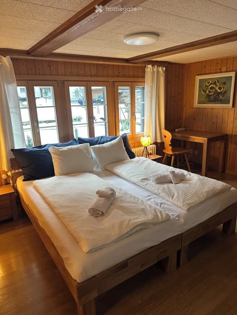 Cozy wooden bedroom with a large bed, nightstand, desk, and large windows letting in natural light.