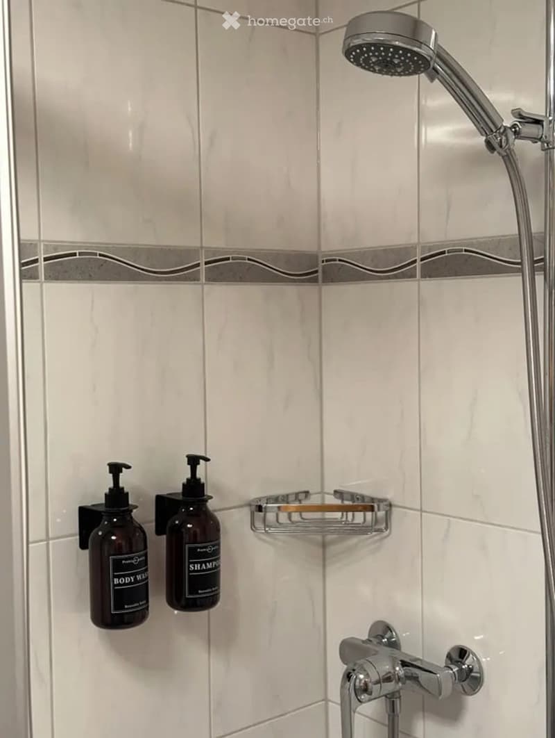 Modern bathroom shower area with white tiles, a handheld showerhead, a chrome faucet, and two bottles of shampoo and body wash on a wall-mounted shelf.