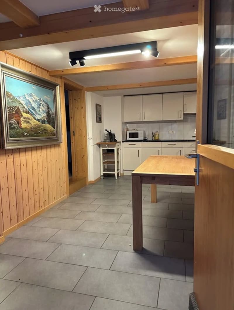The image shows a cozy, well-lit room with wooden panel walls and a tiled floor. A large wooden table is positioned in the foreground, and a small kitchen area is visible in the background, featuring white cabinets, a microwave, and various kitchen items.