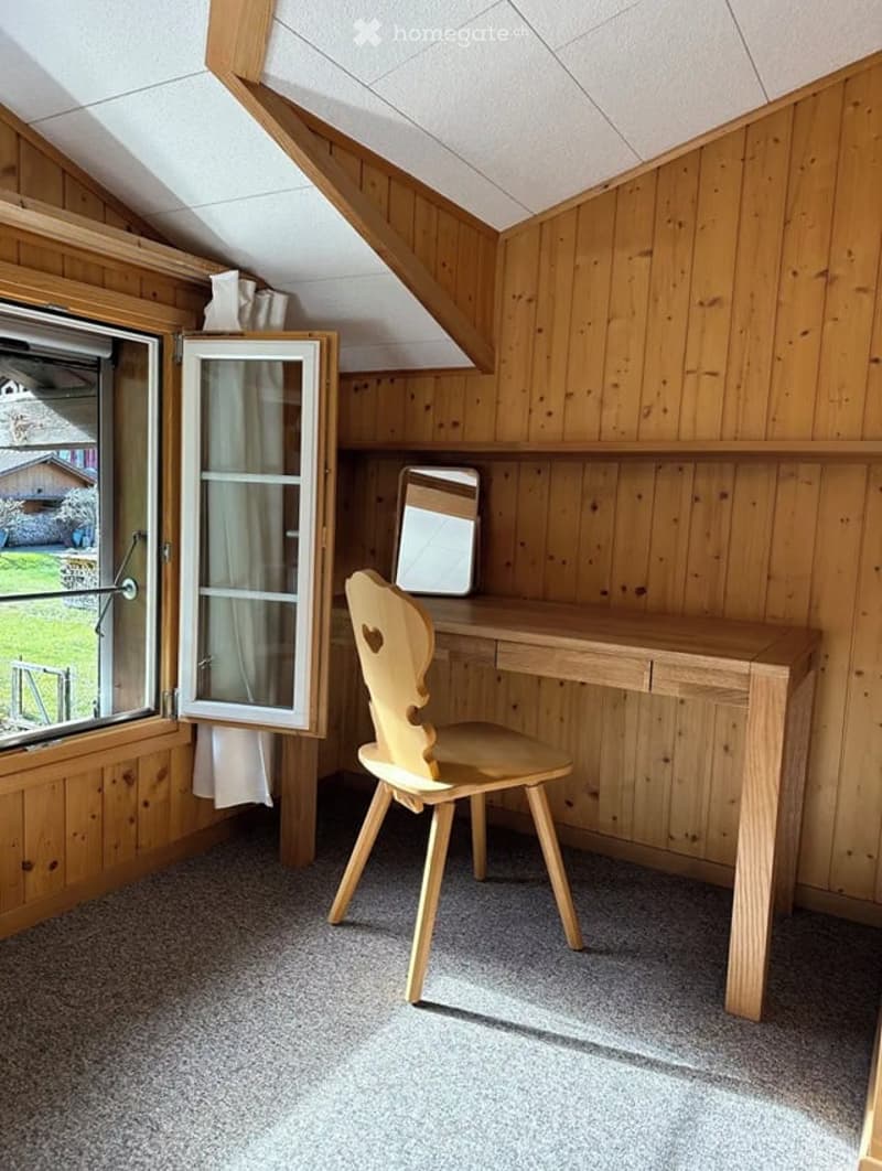 Cozy wooden-paneled room with a slanted ceiling, featuring a wooden chair, desk, mirror, and cabinet with glass doors.
