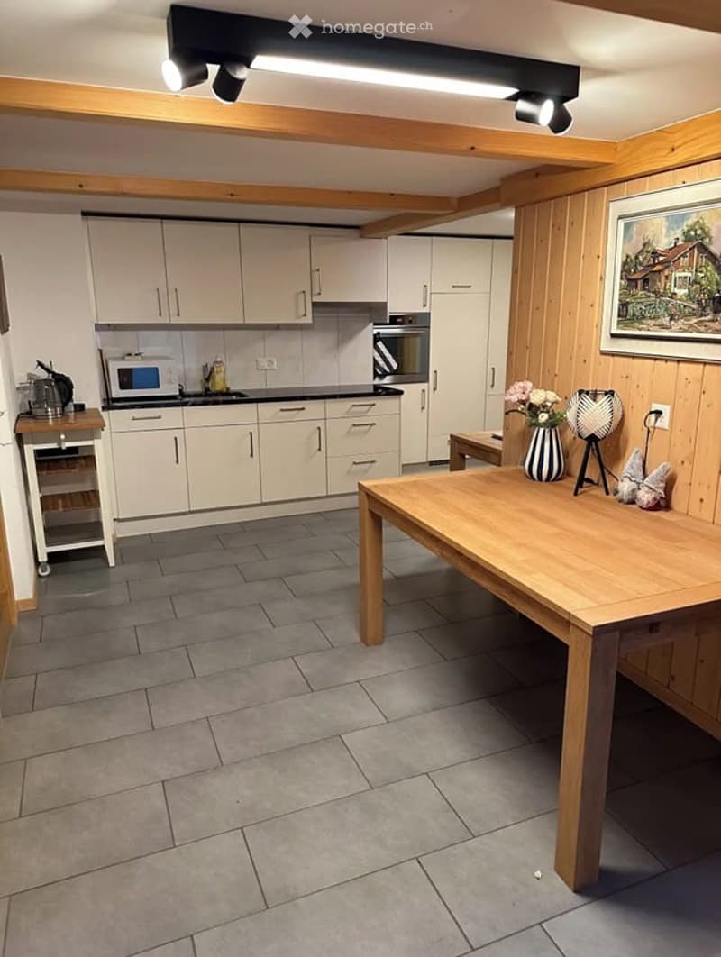 Bright kitchen with white cabinets, grey tiled floor, and wooden beams on the ceiling. Includes a microwave, oven, dining table, chairs, vase with flowers, lamp, and picture frame.
