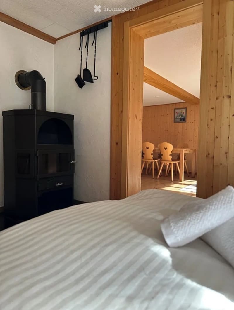Cozy bedroom with a bed, wooden furniture, and a small dining area visible through an open doorway.
