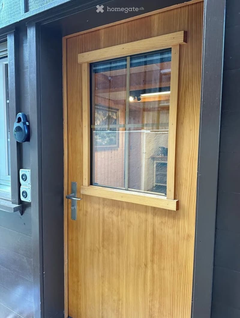 Wooden door with a small window, featuring a silver handle and a security camera mounted on the wall nearby.