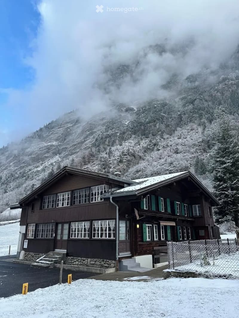A two-story wooden building with green shutters and large windows, situated in a snowy mountainous area with a stone foundation and a fenced perimeter.