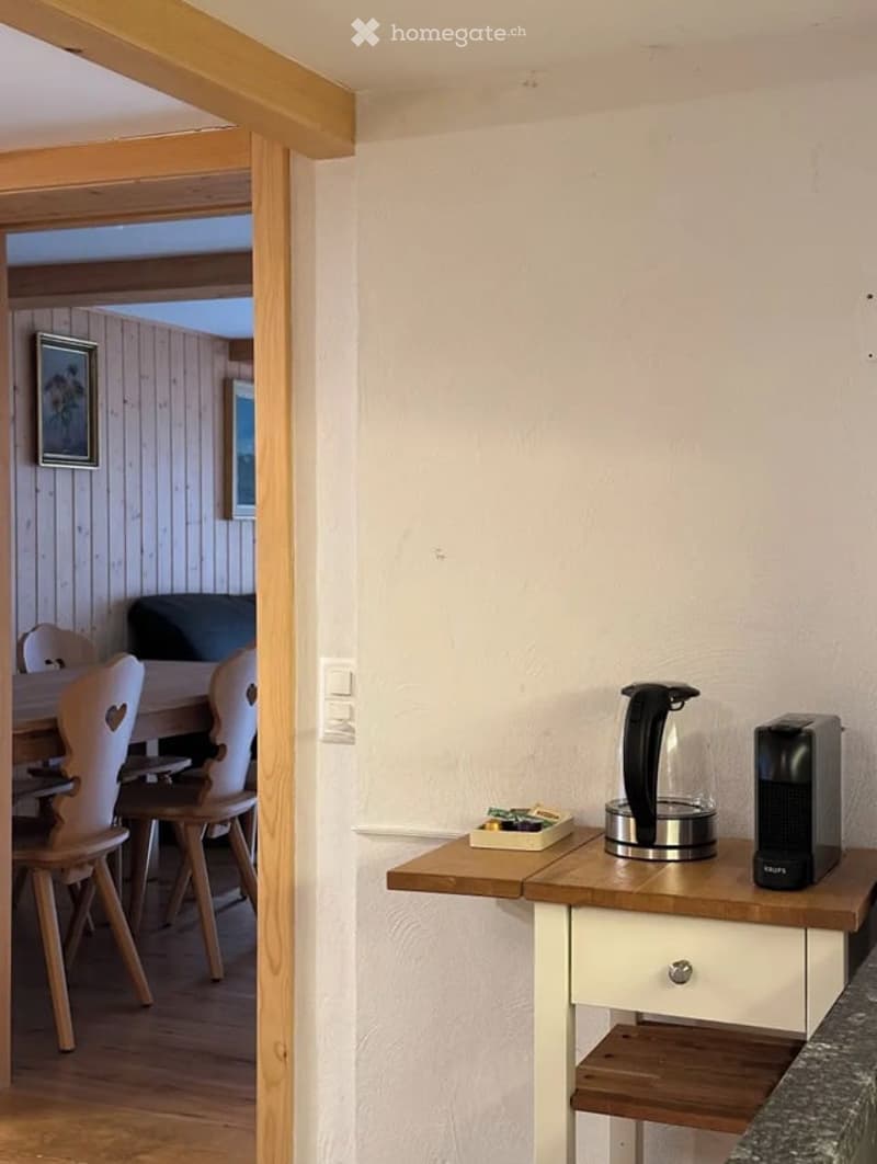 A small, modern kitchen area with a wooden table, white chairs, and a countertop holding a kettle and coffee maker. The adjacent dining area features a wooden dining table and chairs.