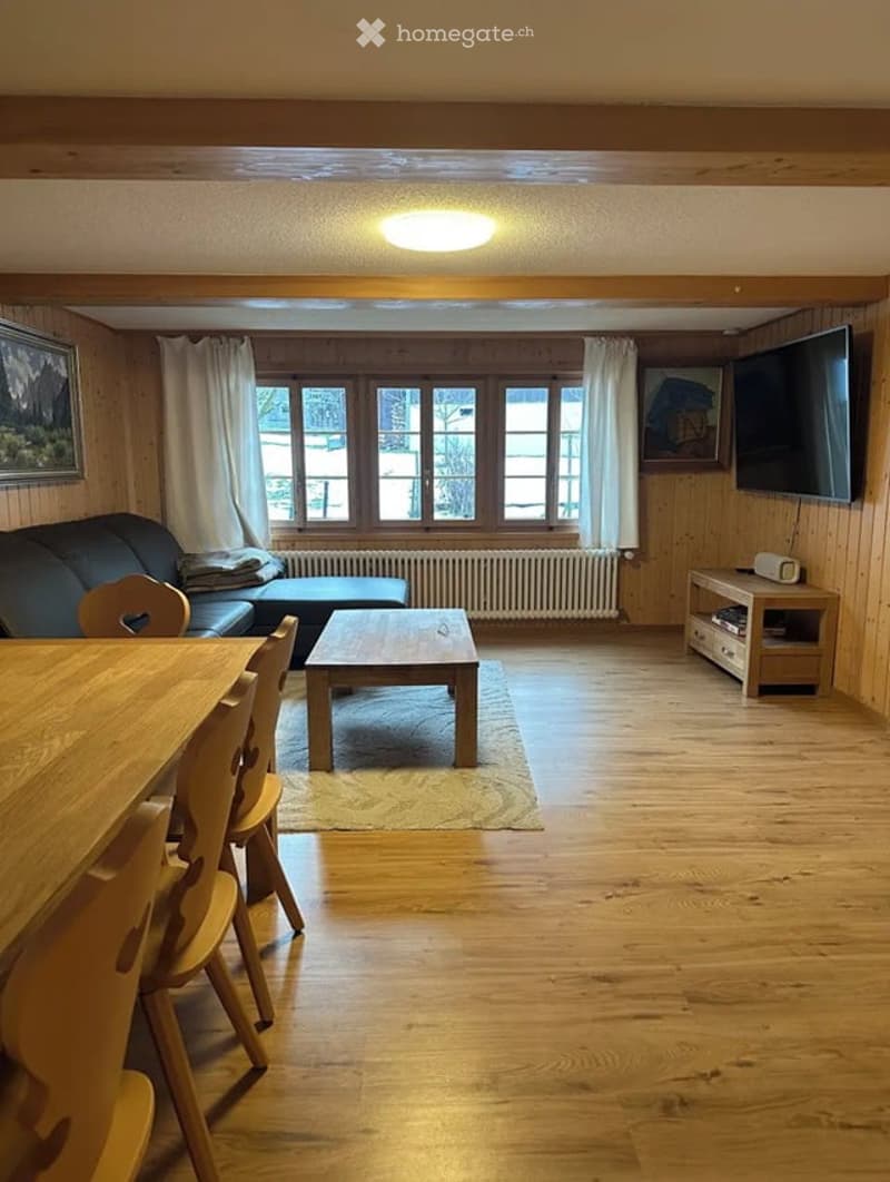 Cozy living and dining area with wooden walls, hardwood floor, large windows, sofa, coffee table, dining table, chairs, and TV stand.