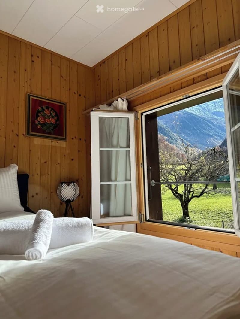 Cozy wooden bedroom with a large window offering a scenic mountain view, featuring a bed with white linens, a decorative lamp, and a framed picture on the wall.