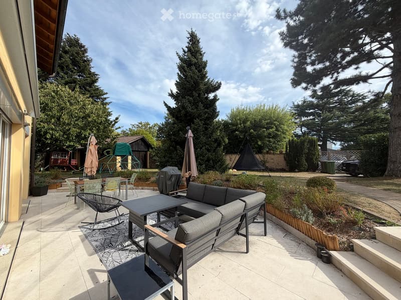 Outdoor patio with a grey sectional sofa, black coffee table, chairs, and umbrellas, surrounded by greenery and trees.