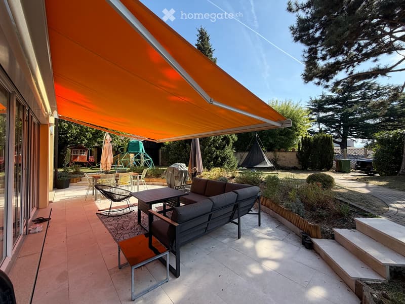 Outdoor patio with a covered awning, sofa, table, chairs, and umbrella, surrounded by greenery and steps leading to a garden area.