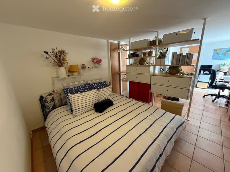 Cozy bedroom with a single bed, striped bedding, nightstand, shelves with decorative items, and an adjacent workspace area with a desk and office chair.