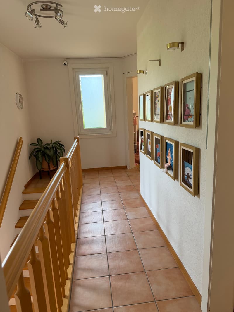 Hallway with tiled floor, wooden staircase railing, framed artwork on walls, and a potted plant near the stairs.