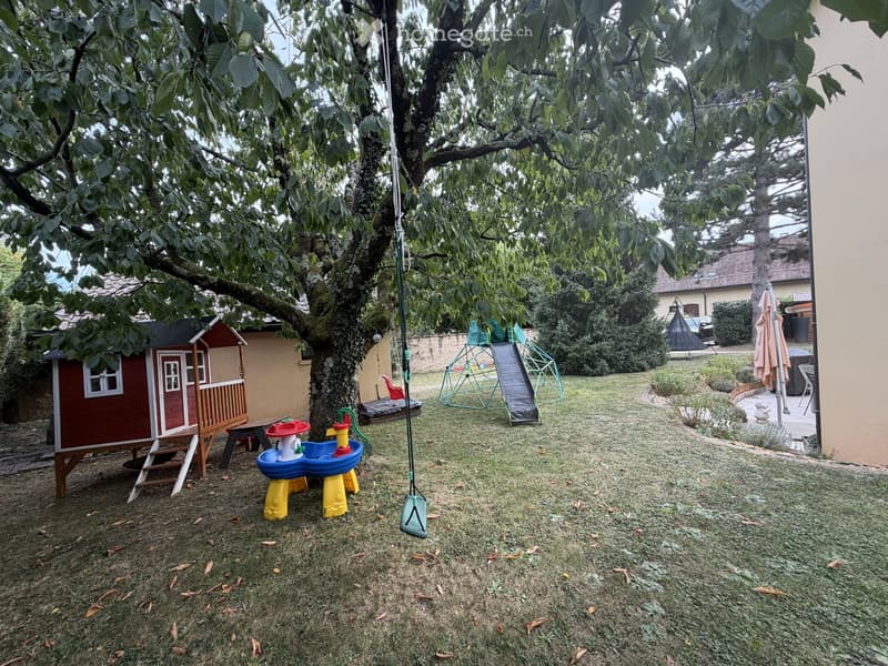 A well-maintained garden with a swing, a small playhouse, a colorful children's fountain, a slide, and a climbing frame. The area is surrounded by trees and greenery, with a house visible in the background.